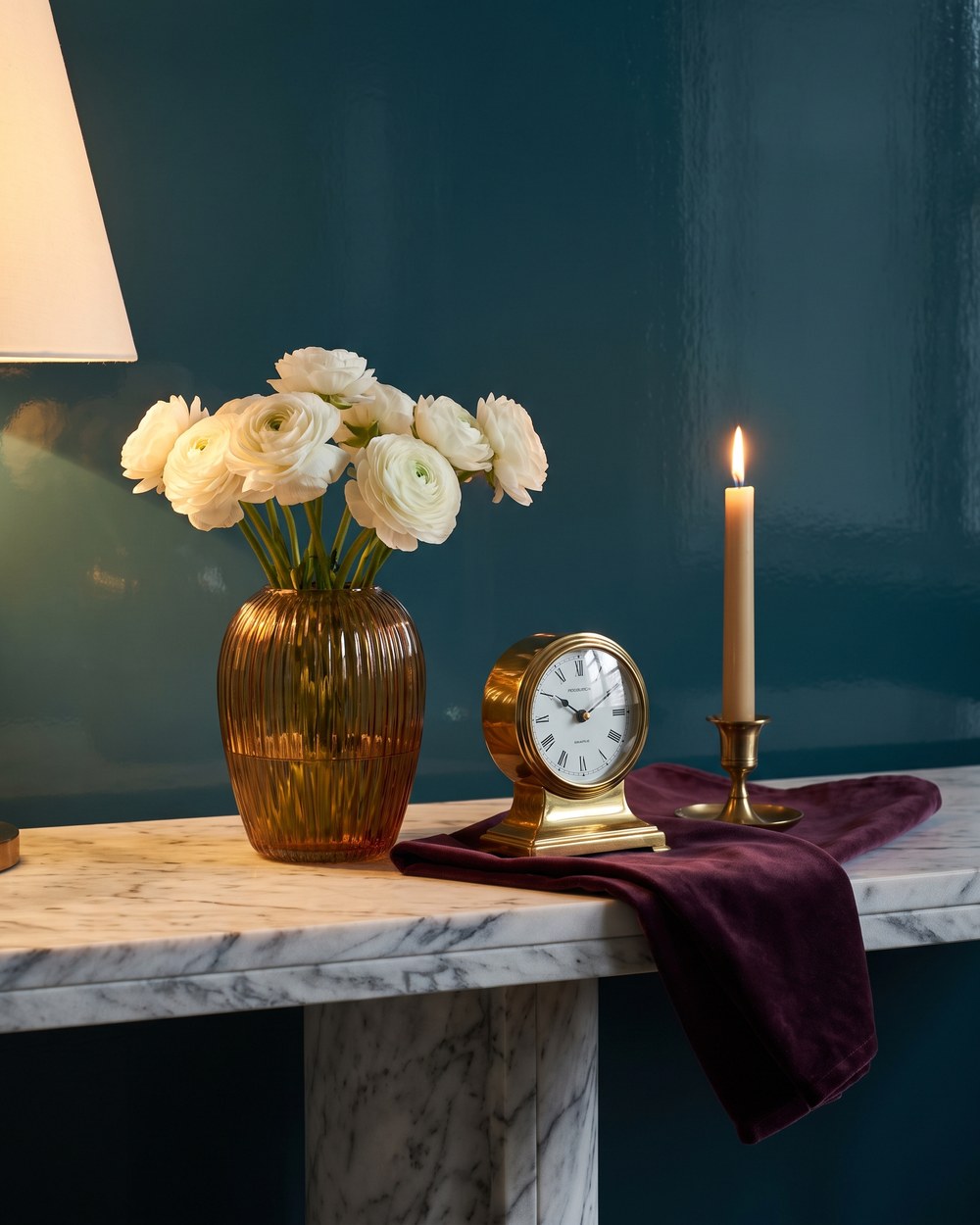 A marble console in the reception room holding a ribbed amber vase of white ranunculus, a small brass clock and a lit candle